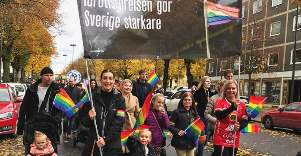 Stor folkskara går i ett pridetåg med prideflaggor och håller upp en skylt som säger "Idrottsrörelsen gör Sverige starkare"
