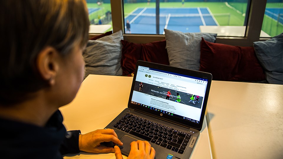 Kvinna sitter med dator
ovanför en tennisplan. 
