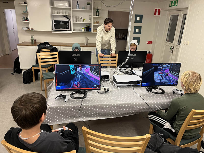 Bord med dataspelsskärmar och en grupp barn som sitter och spelar. En ledare står upp och tittar på.
