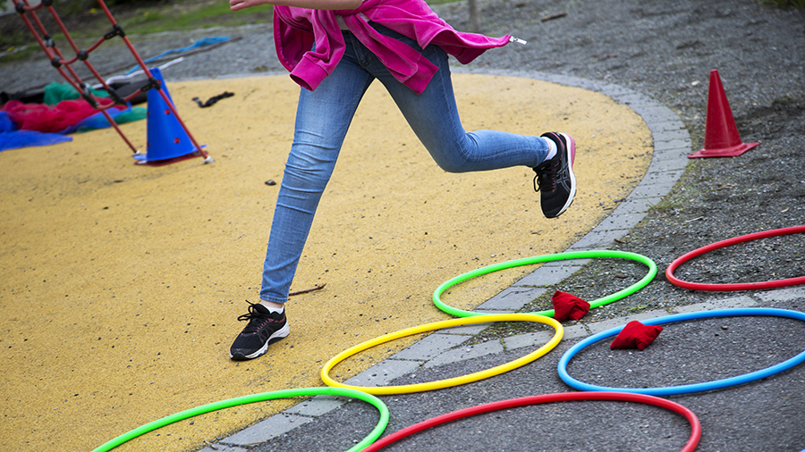 Ett par ben som hoppar över och i färgglada ringar som ligger på marken, i en rörelsebana, på en skolgård.